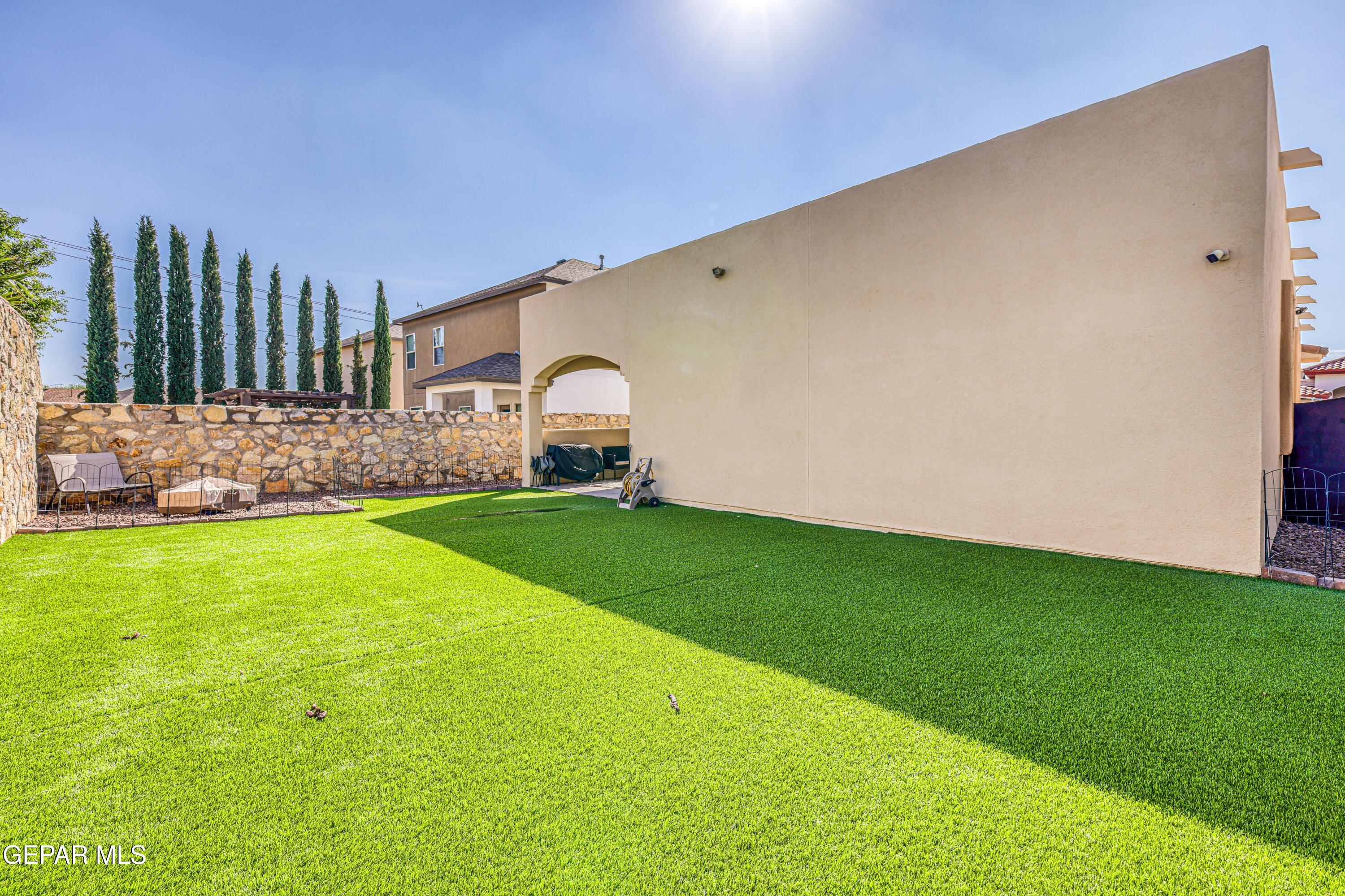 Backyard with a broad green lawn, stone wall, and shaded patio beside a tan stucco home.