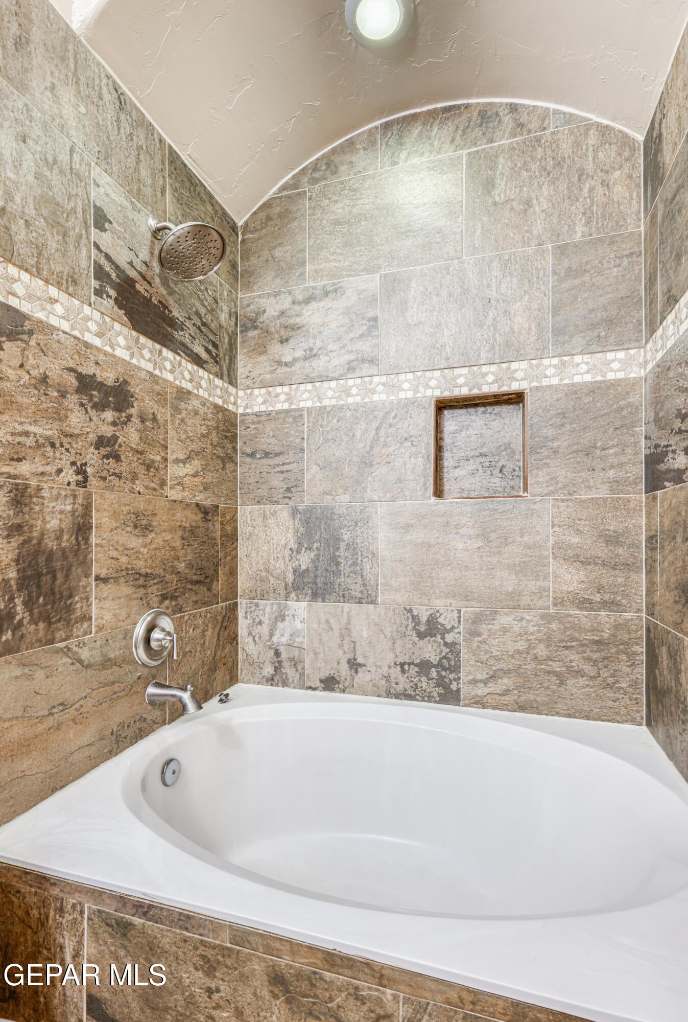 White soaking tub beneath a tiled shower with a decorative mosaic band and built-in niche.