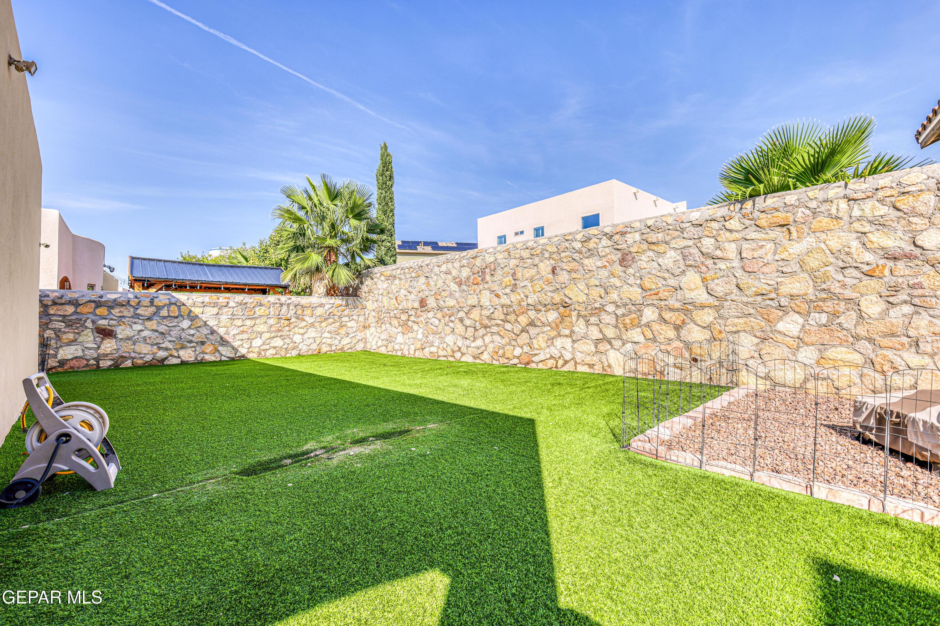 Backyard with green lawn, high stone walls, palm trees, and a fenced gravel garden bed.
