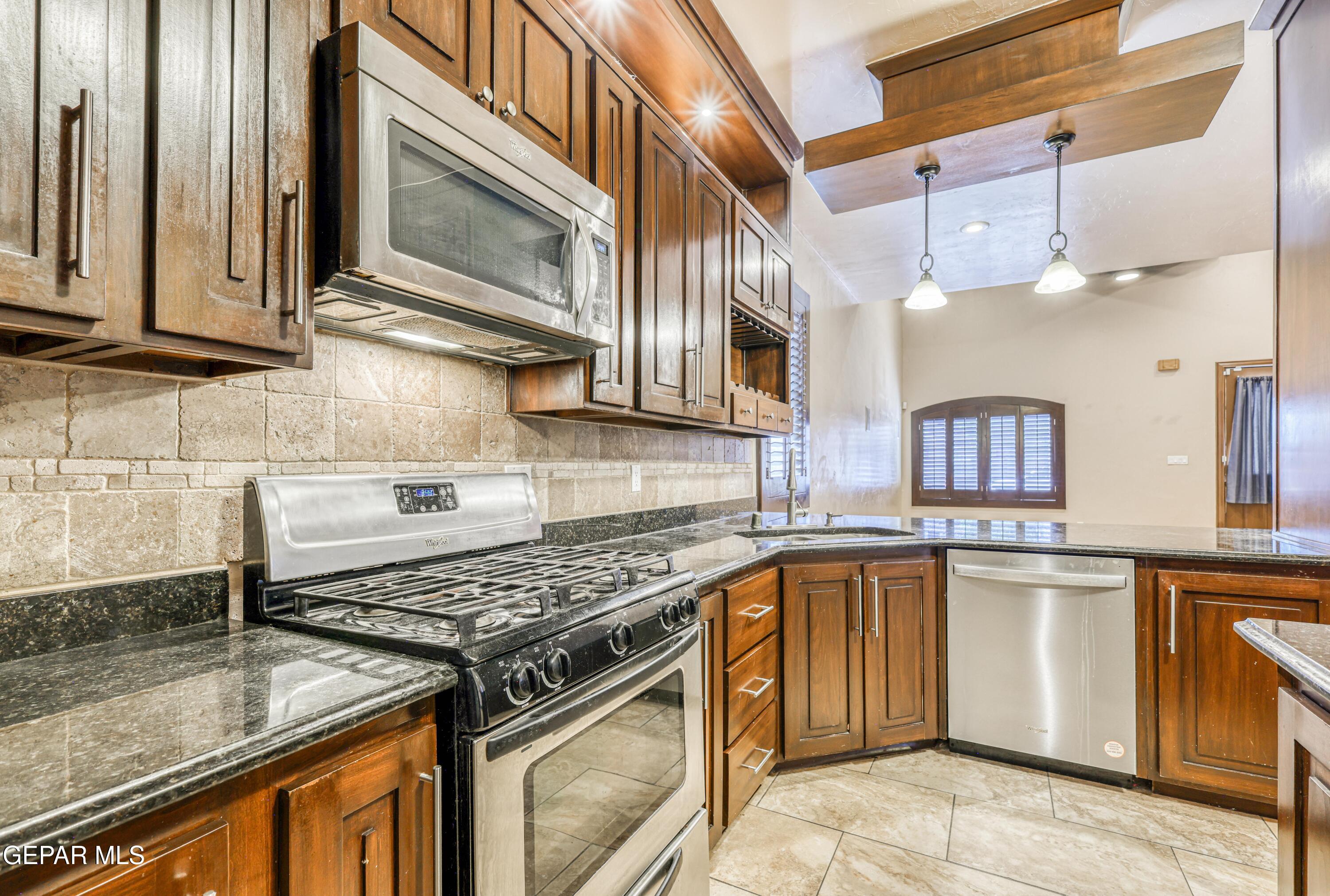 Kitchen with dark wood cabinets, stone backsplash, stainless steel range and dishwasher, and pendant lights.