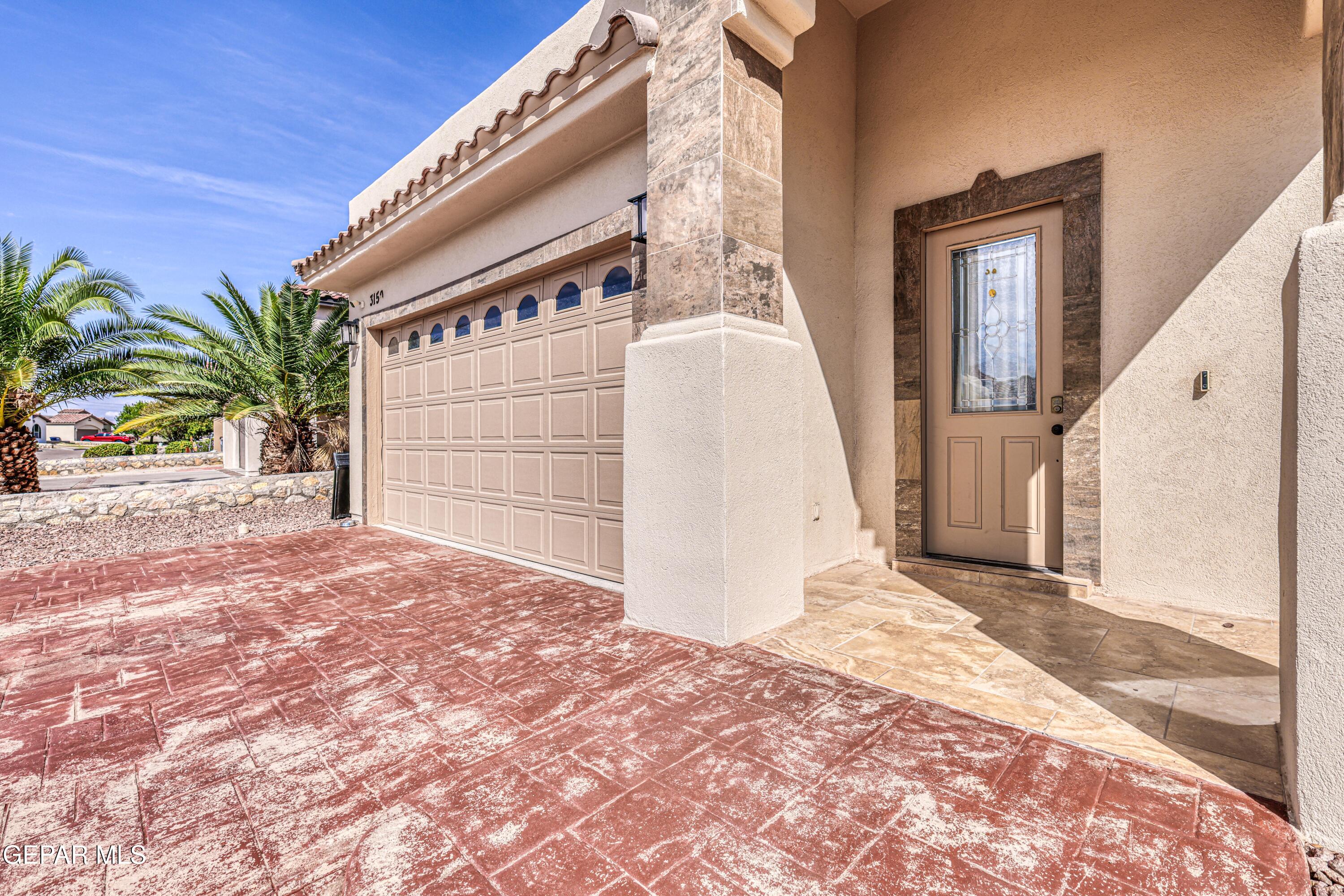 Stucco home entrance and garage with a decorative-glass door, stone trim, and red stamped-concrete driveway.