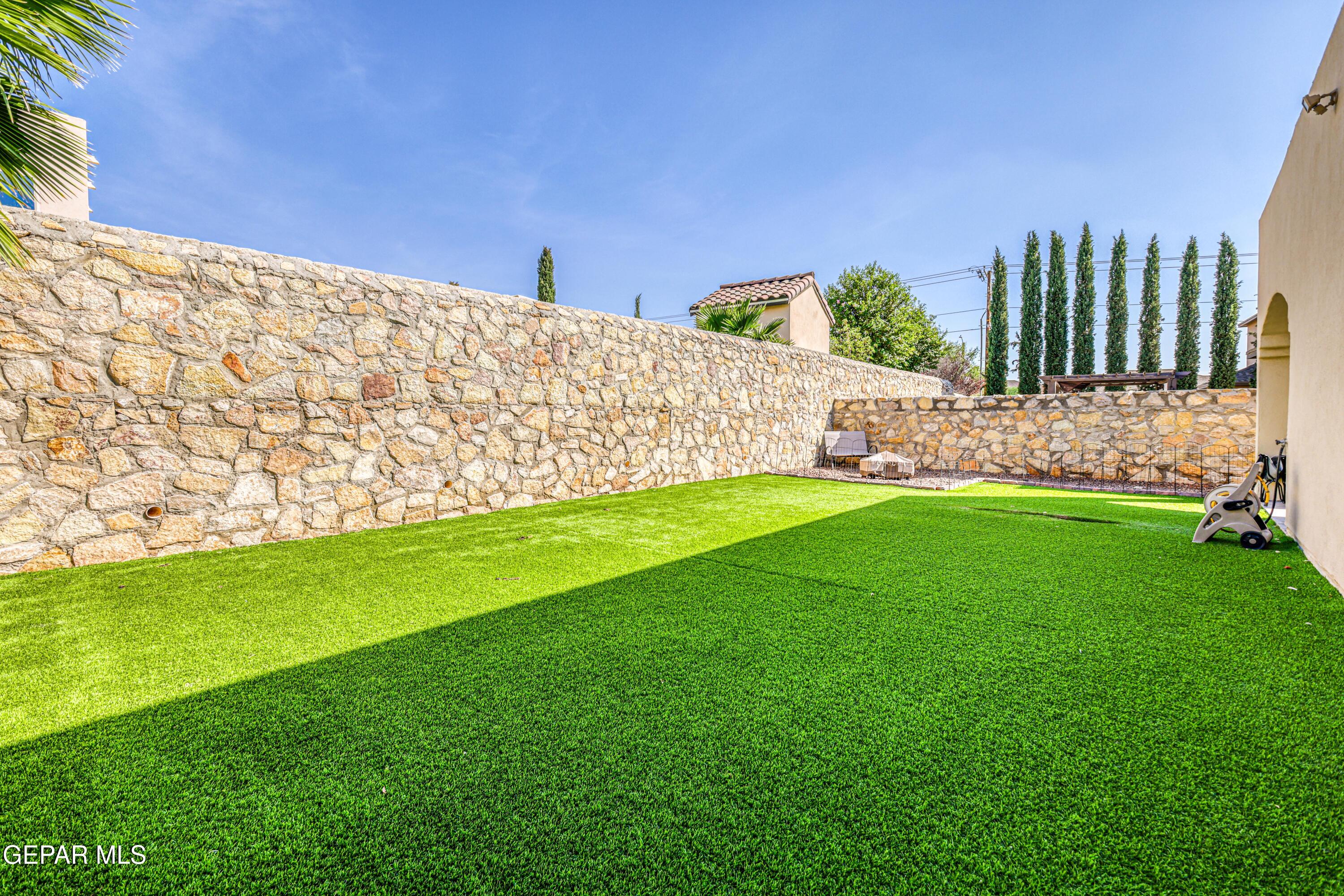 Backyard with a broad green lawn, tall stone walls, and slender evergreen trees.