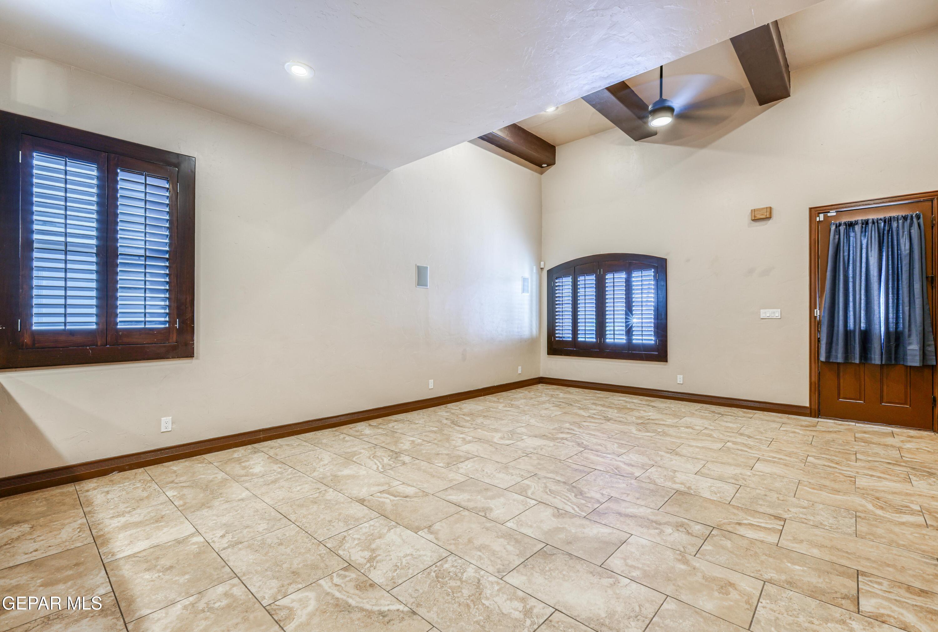 Empty tiled room with dark wood shuttered windows, exposed ceiling beams, and a ceiling fan.