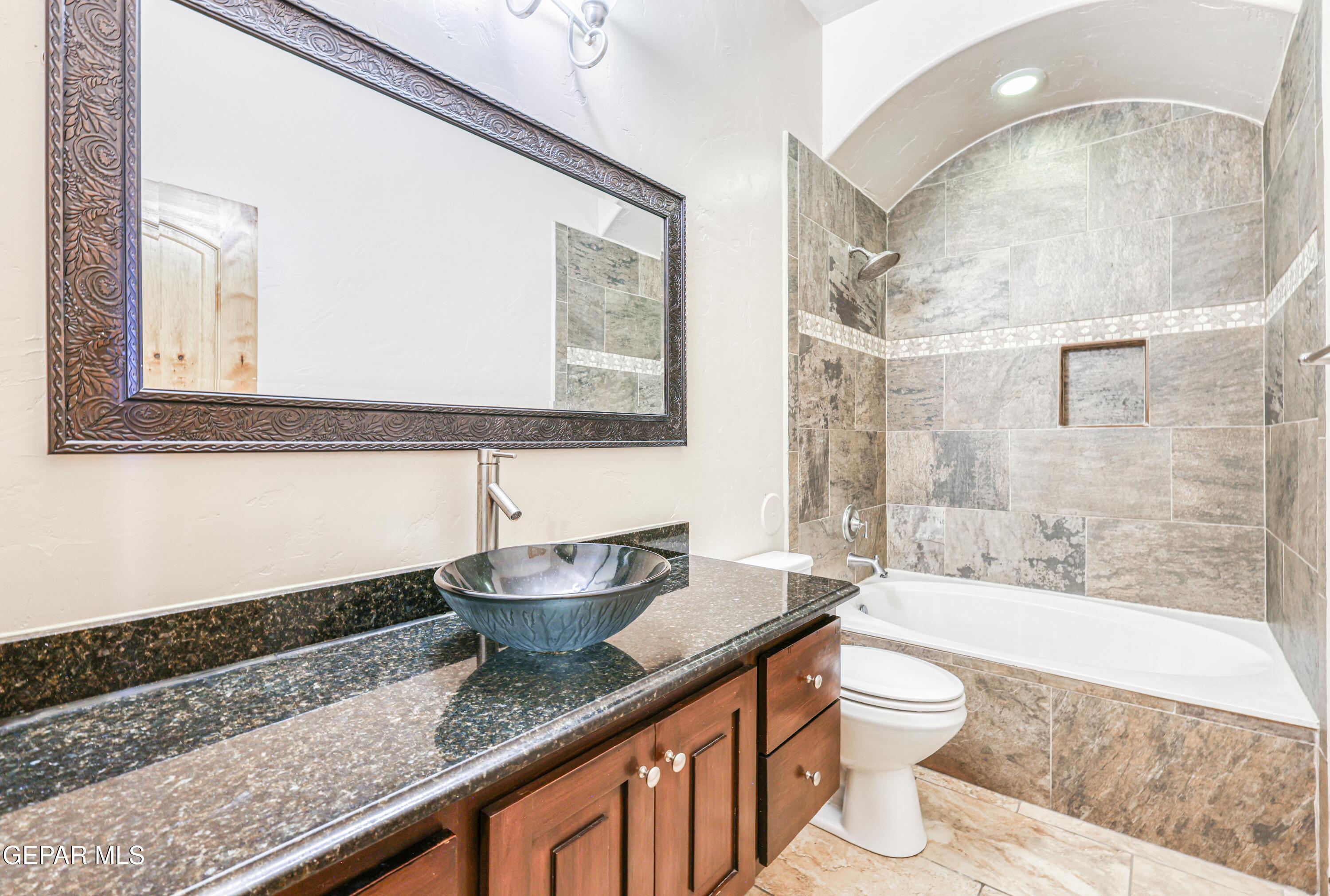 Bathroom with a granite vanity, glass vessel sink, and tiled tub and shower beneath an arched ceiling.