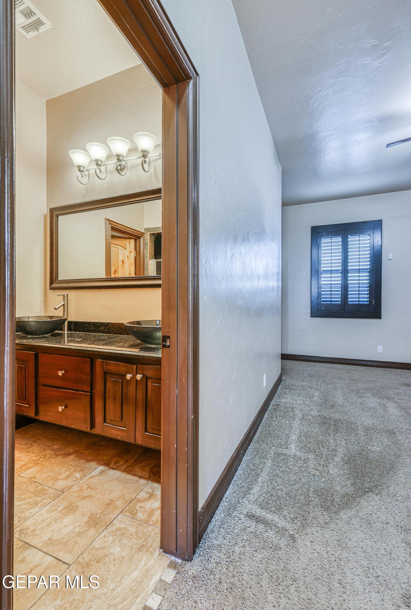 Bathroom vanity with dual vessel sinks beside an empty carpeted room with a shuttered window.