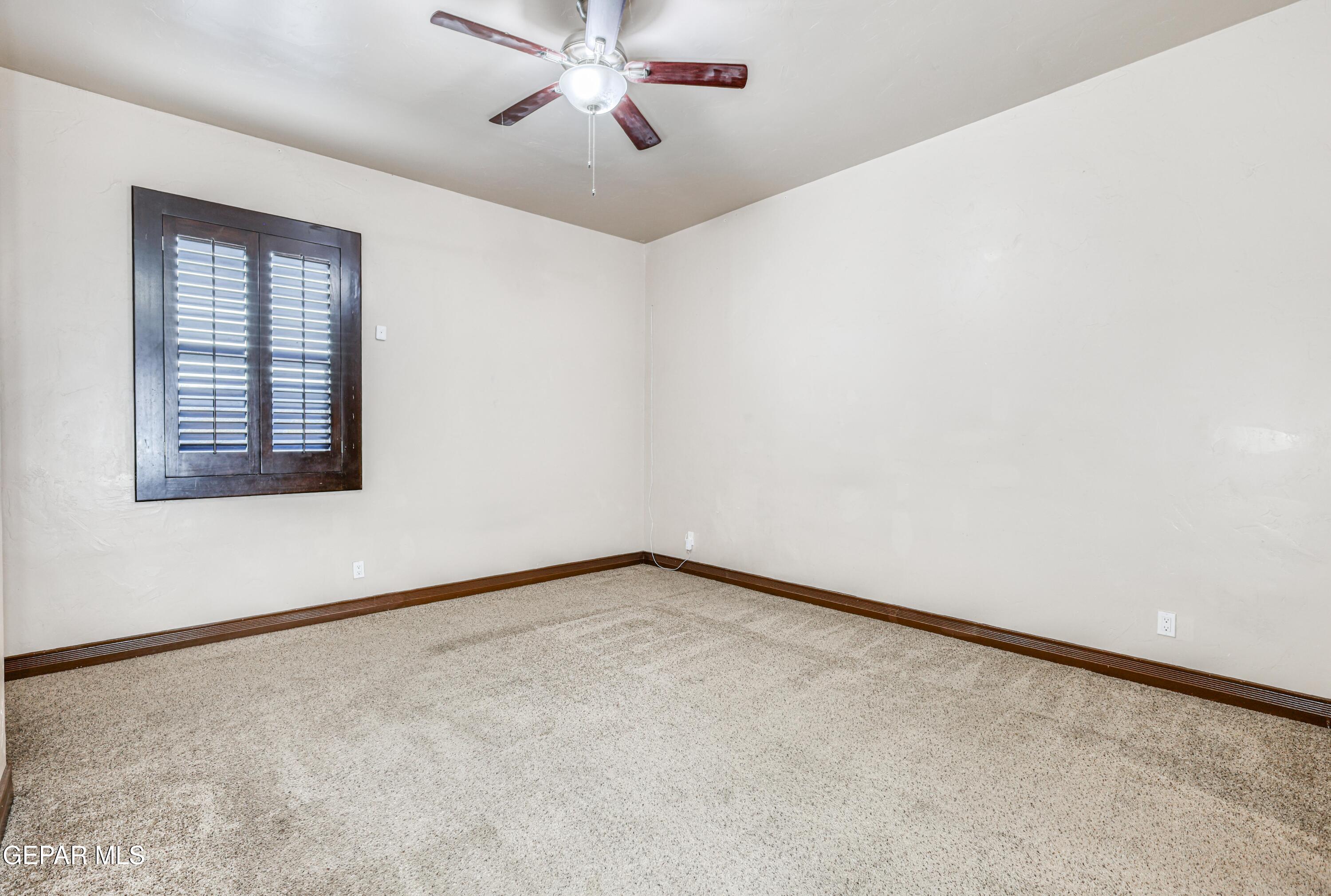 Vacant bedroom with beige carpet, a dark-framed shuttered window, and a ceiling fan.