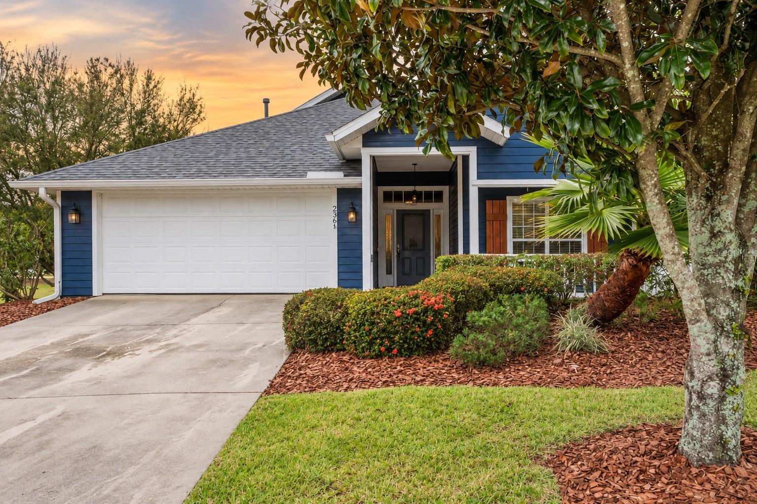 Blue home with a white two-car garage, landscaped front yard, and covered entrance at sunset.