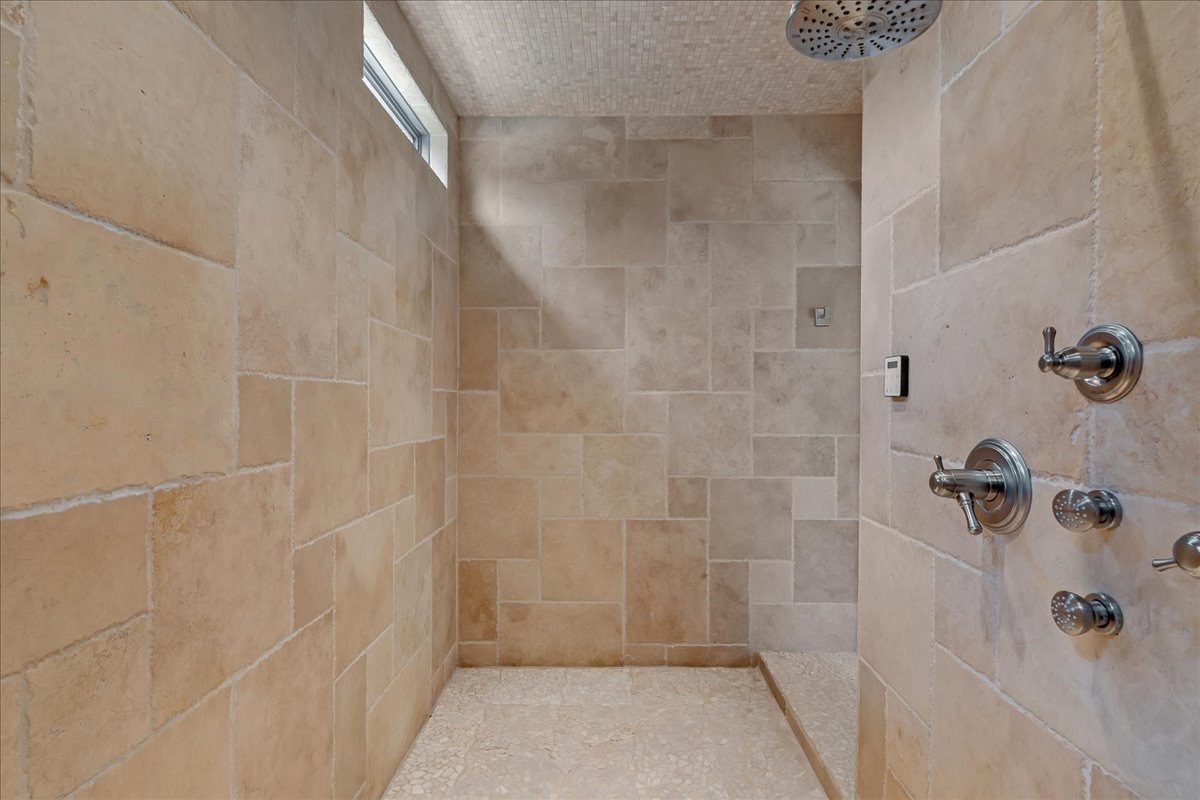 Walk-in shower with beige stone tile, multiple shower fixtures, and a high narrow window.