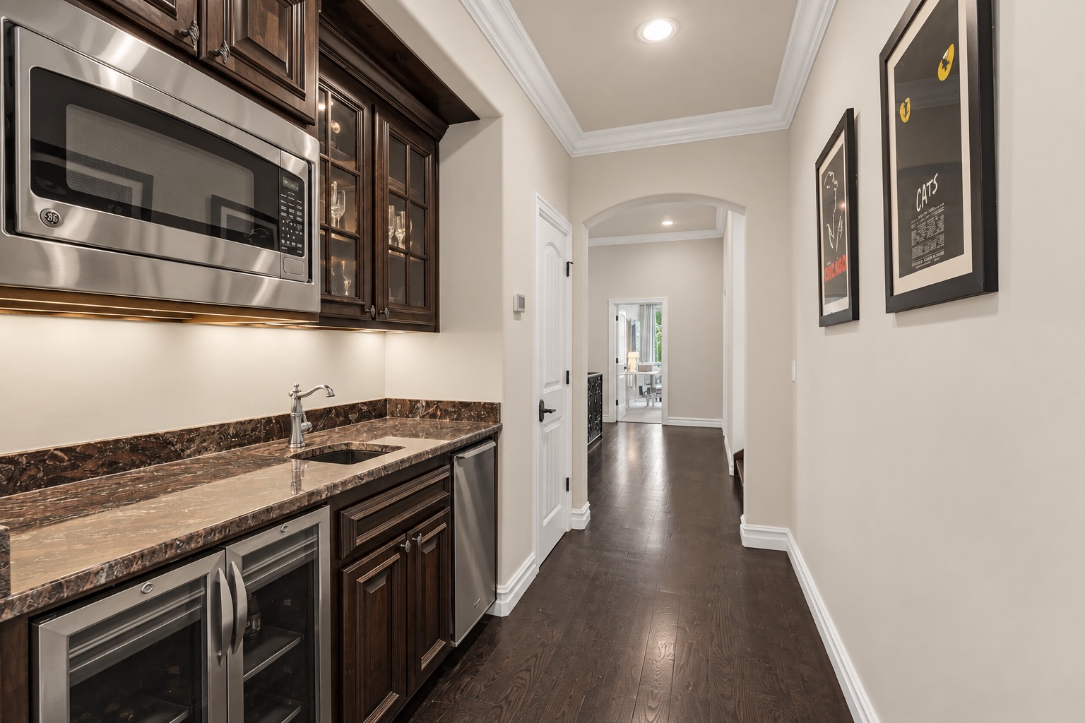 Kitchen with dark wood cabinetry, stone counters, wine refrigerators, and a hallway leading to a bright room.