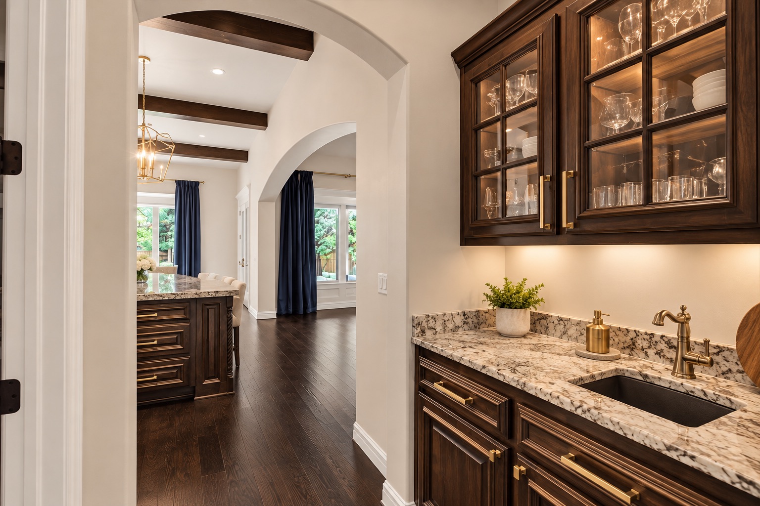 Wet bar with glass-front cabinets, a stone counter, and sink beside an arched dining room entrance.