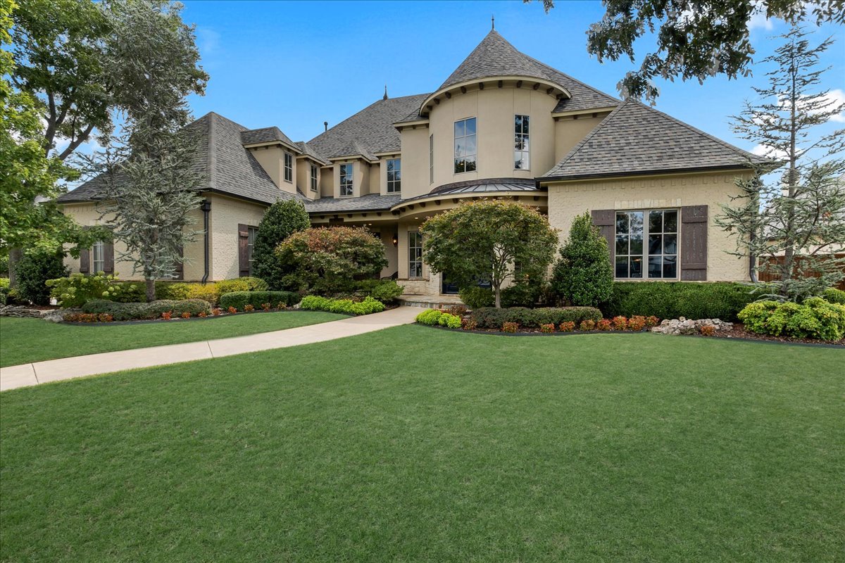 Large beige home with a round turret, landscaped flower beds, and a curved walkway across the lawn.