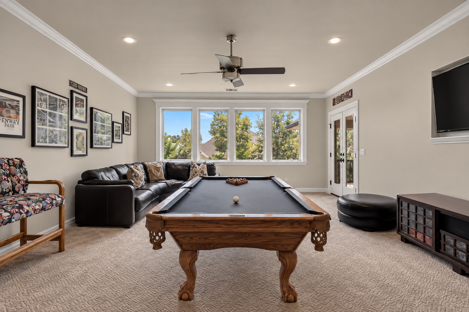 Game room with a wooden pool table, black sectional sofa, and large windows overlooking trees.