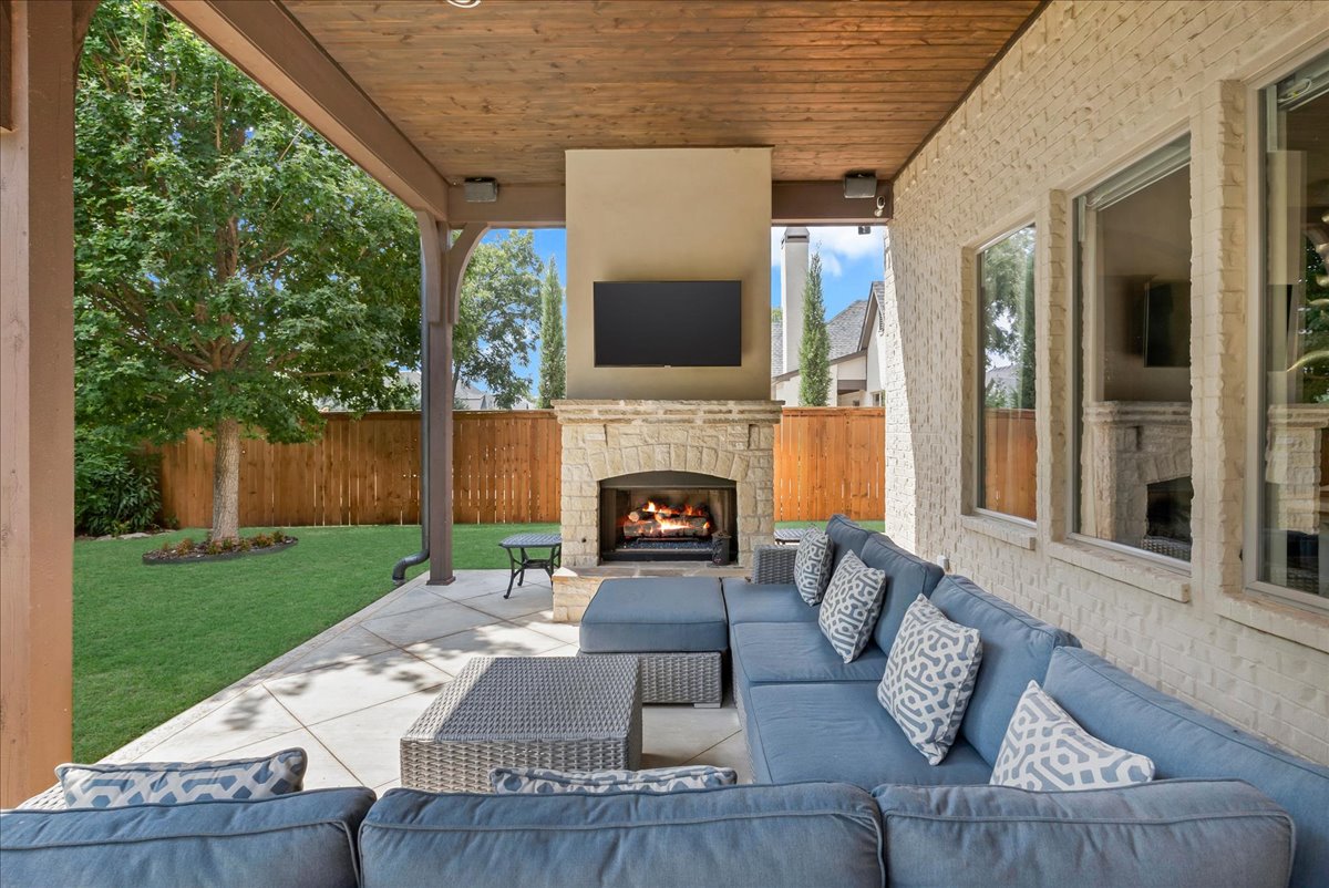 Covered patio with stone fireplace, mounted TV, and blue sectional overlooking a fenced lawn.