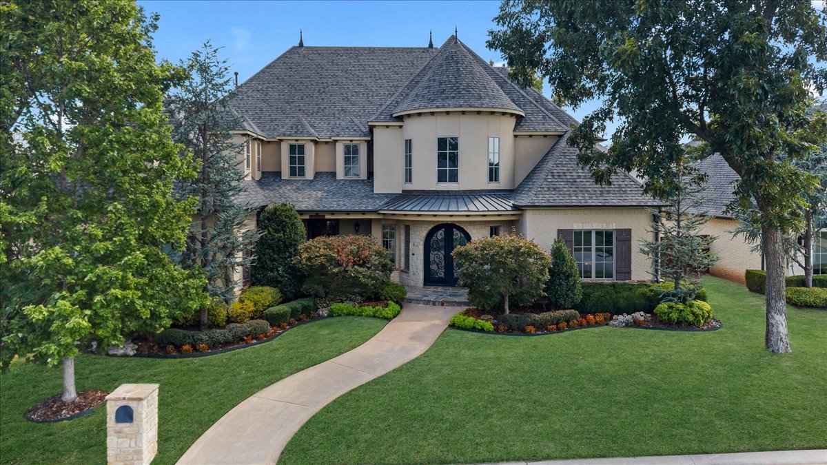 Two-story beige home with a turret-like bay, arched entry, curved walkway, and landscaped front lawn.