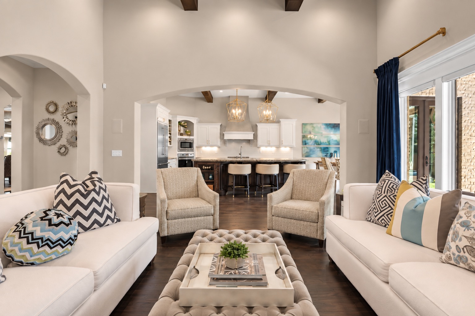 Living room with white sofas and tufted ottoman, opening onto a bright kitchen.