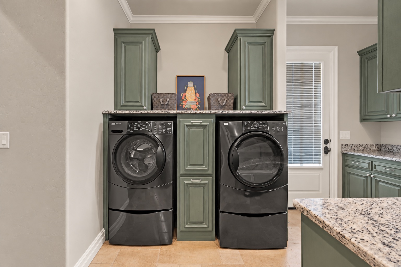Laundry room with dark front-loading washer and dryer beneath green cabinetry and granite countertops.