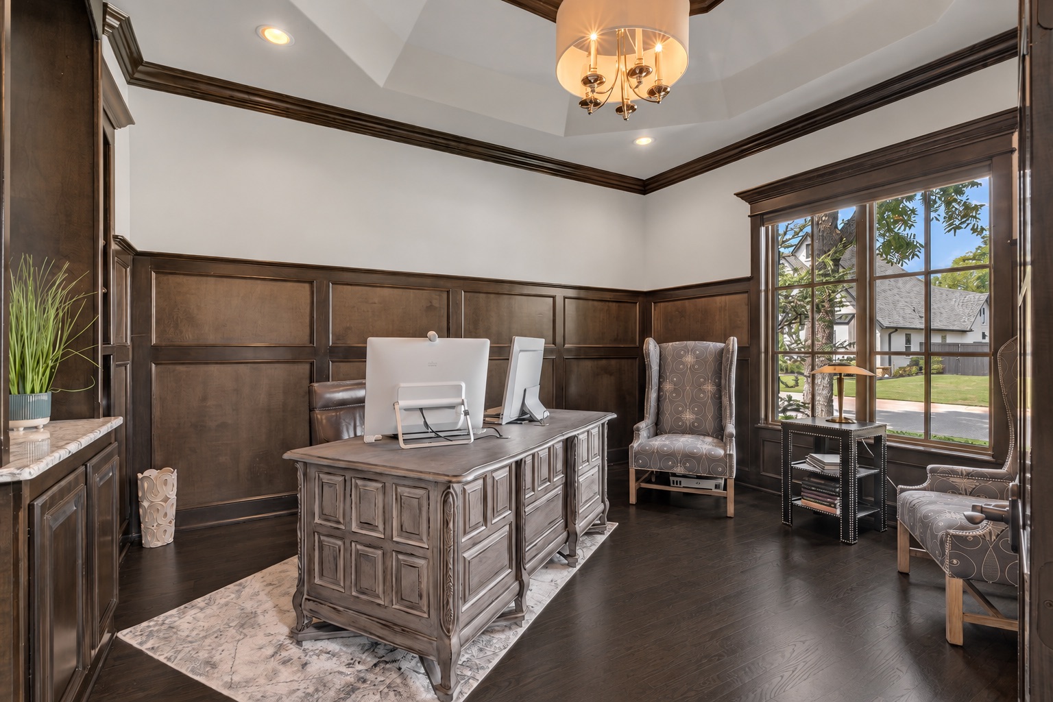Furnished home office with dark wood paneling, a large desk, upholstered chairs, and a broad lawn-facing window.