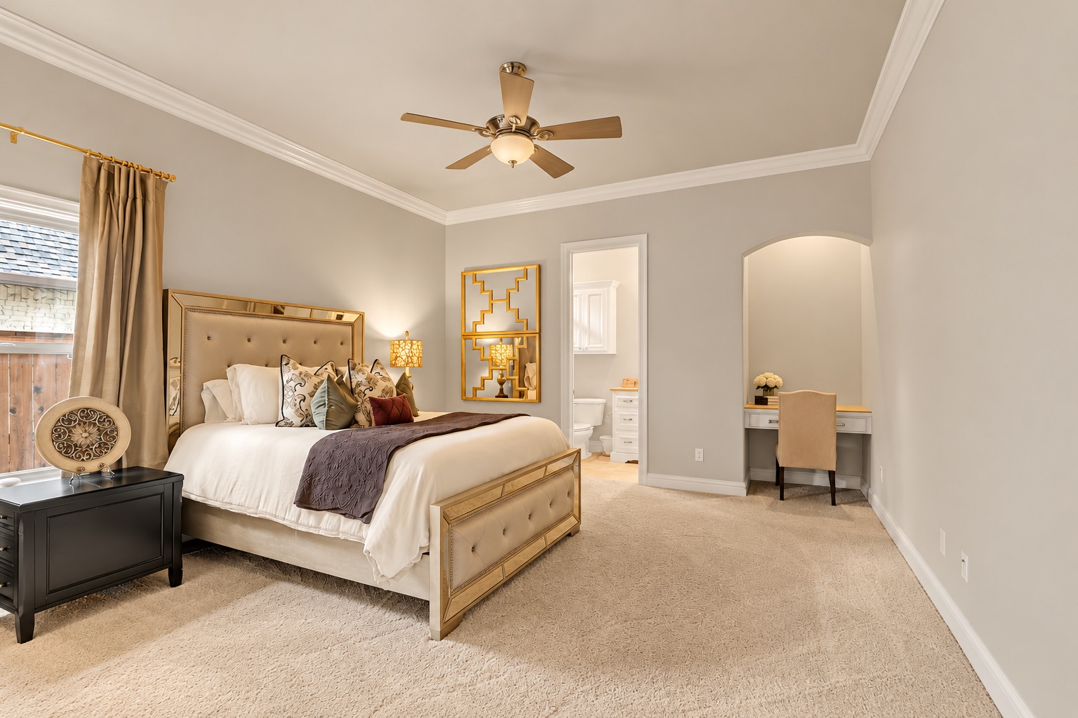 Staged bedroom with a large upholstered bed, ceiling fan, ensuite doorway, and compact desk nook.