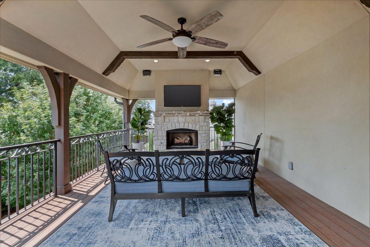 Covered patio with stone fireplace, mounted television, and black metal seating overlooking leafy trees.