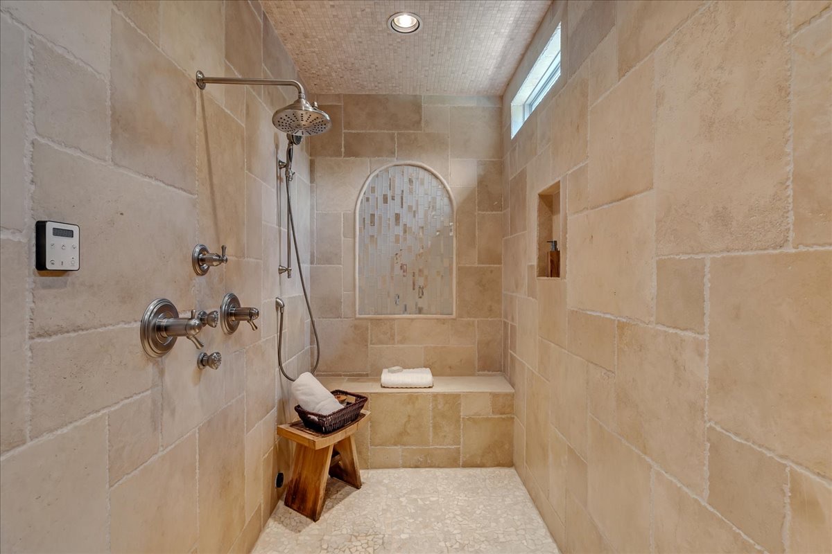 Walk-in shower with beige stone tile, rainfall showerhead, built-in bench, and arched mosaic niche.