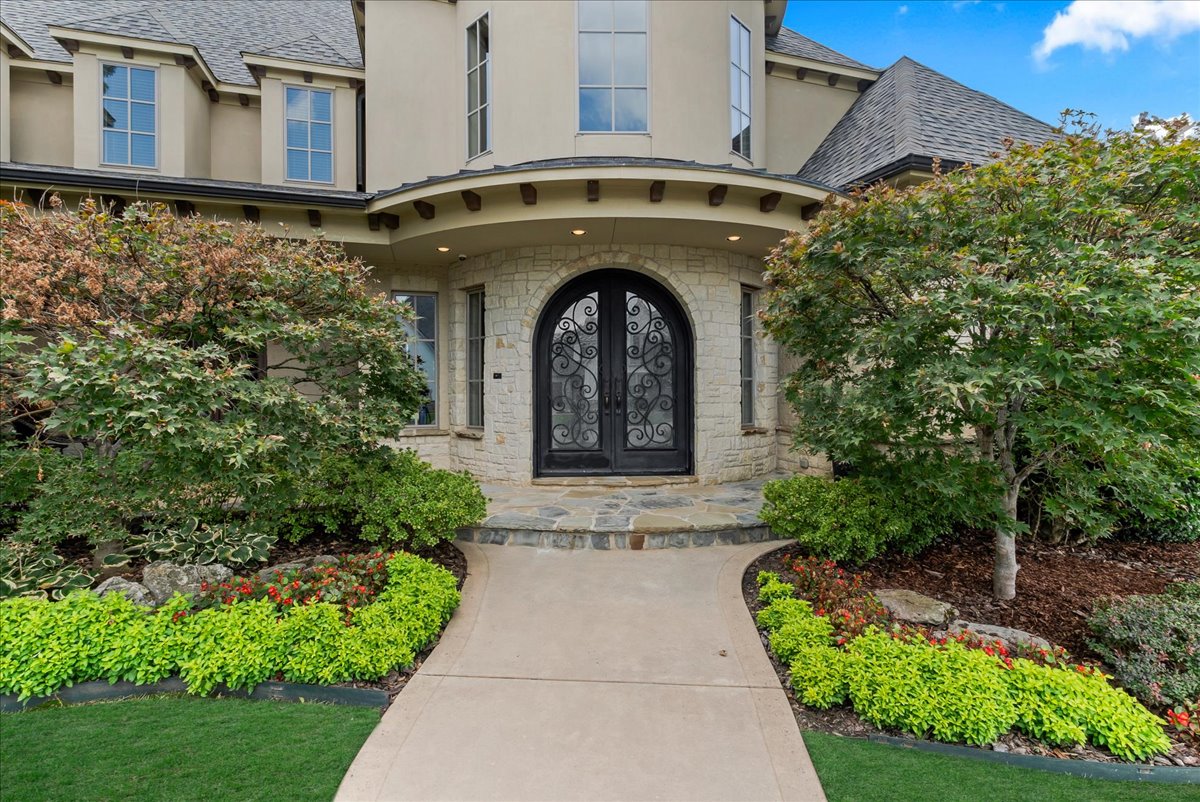 Two-story beige house entrance with ornate black doors and a landscaped curved walkway.