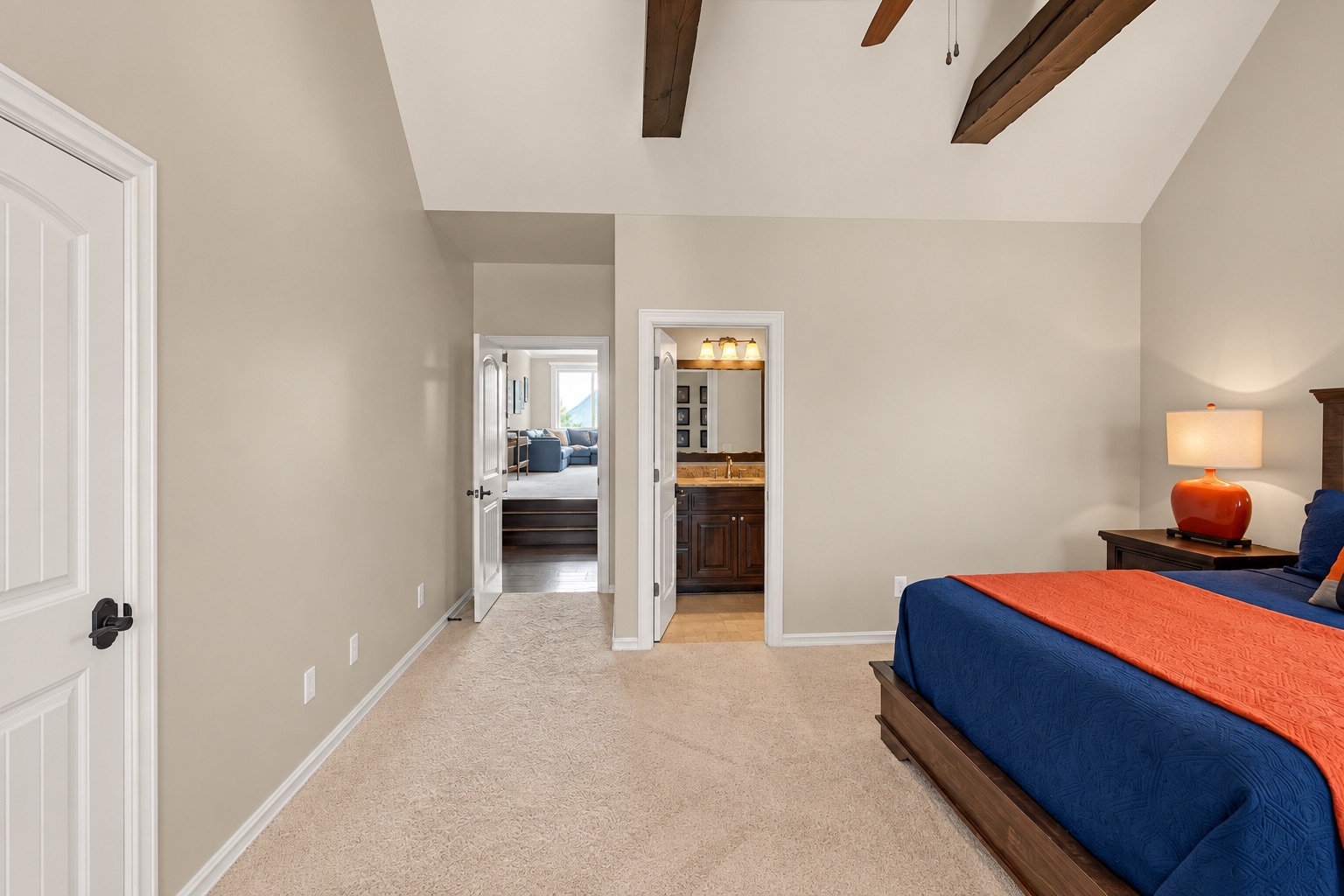 Bedroom with vaulted wood-beam ceiling, blue bed, orange coverlet, and doorway to a bathroom.