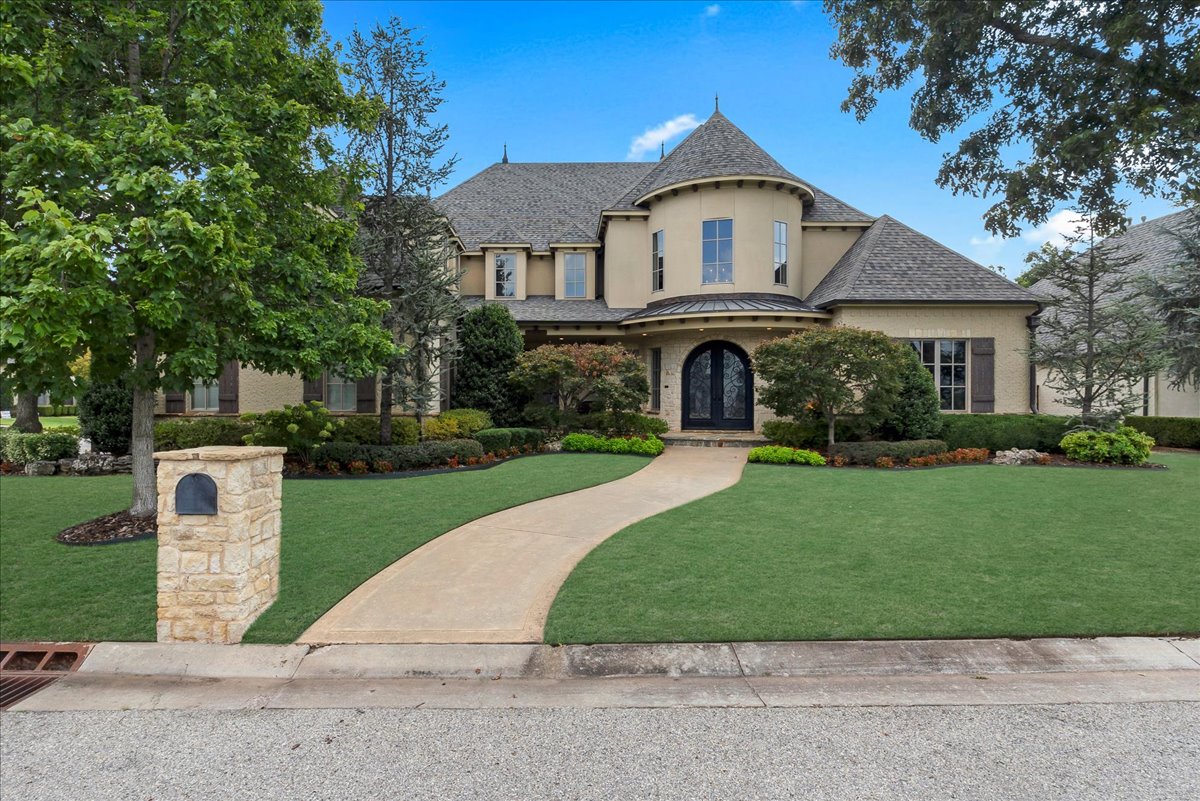 Two-story beige home with a turret-like bay, arched entry, curved walkway, and landscaped lawn.