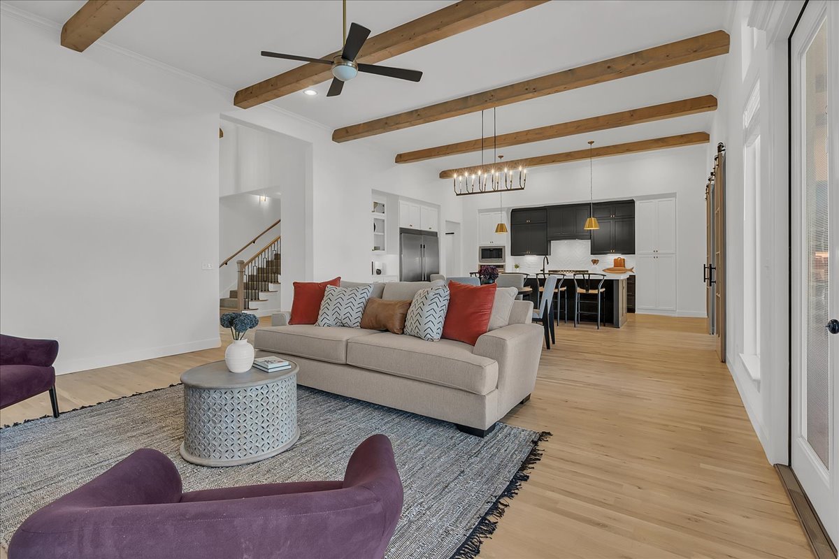 Open living room with exposed wood ceiling beams, neutral sofa, purple chairs, and kitchen beyond.