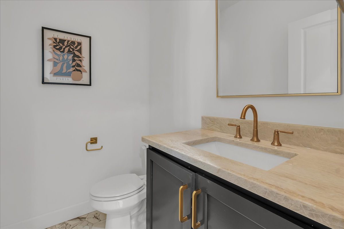 Bathroom with a dark gray vanity, beige stone counter, brass fixtures, and a white toilet.