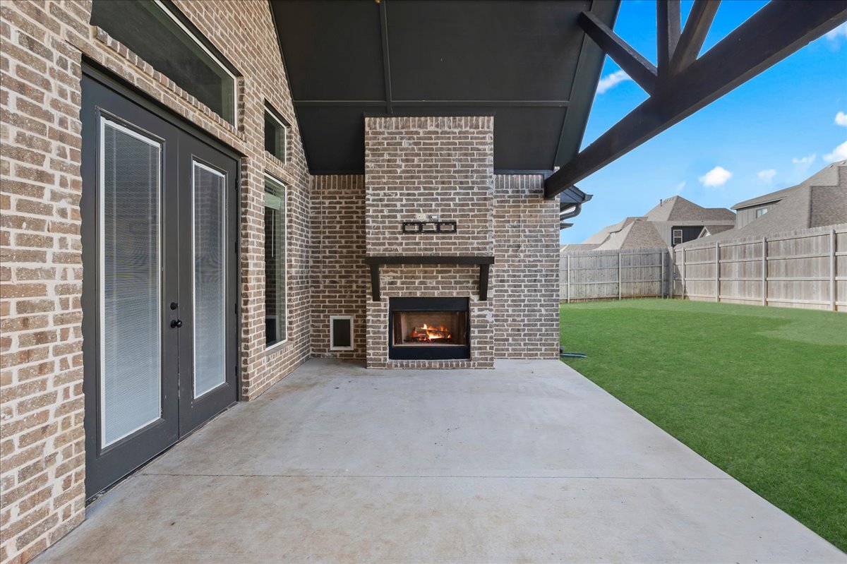 Covered brick patio with a lit fireplace, glass doors, and fenced lawn.