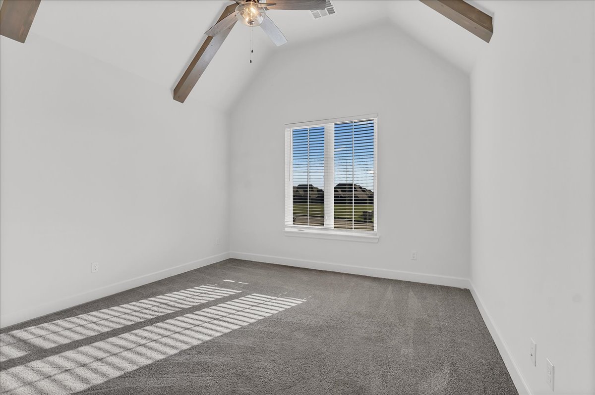 Empty bedroom with gray carpet, exposed ceiling beams, and sunlight through window blinds.