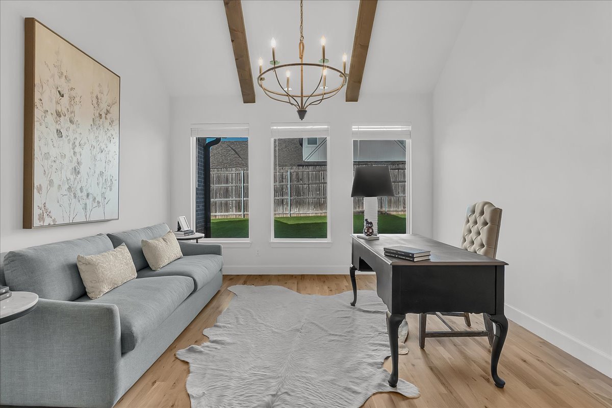 Staged home office with a dark desk, gray sofa, exposed beams, and windows overlooking a fenced yard.