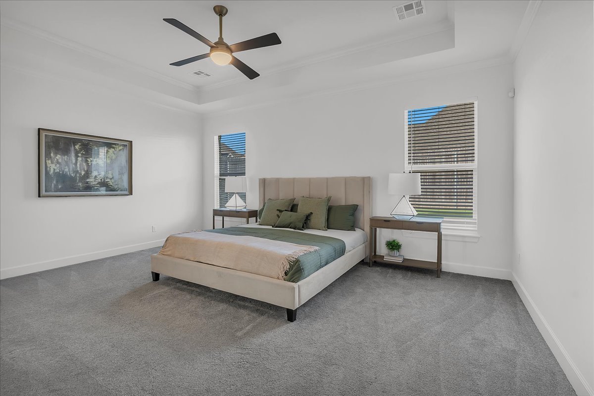 Staged bedroom with a beige upholstered bed, green bedding, gray carpet, and a ceiling fan.