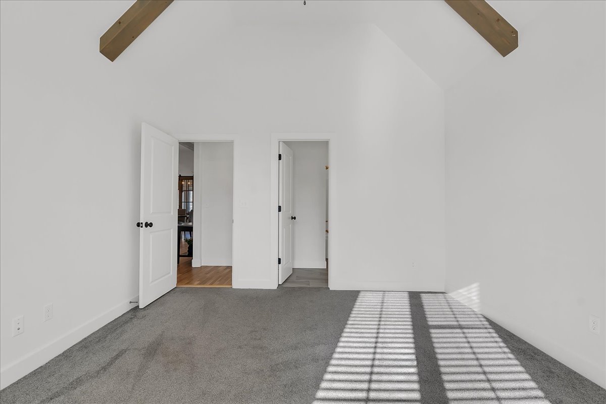 Empty bedroom with vaulted ceiling, exposed beams, gray carpet, and sunlight casting window-grid shadows.