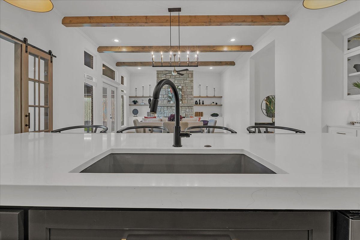 Open kitchen with a white quartz island, black faucet, exposed beams, and stone fireplace beyond.