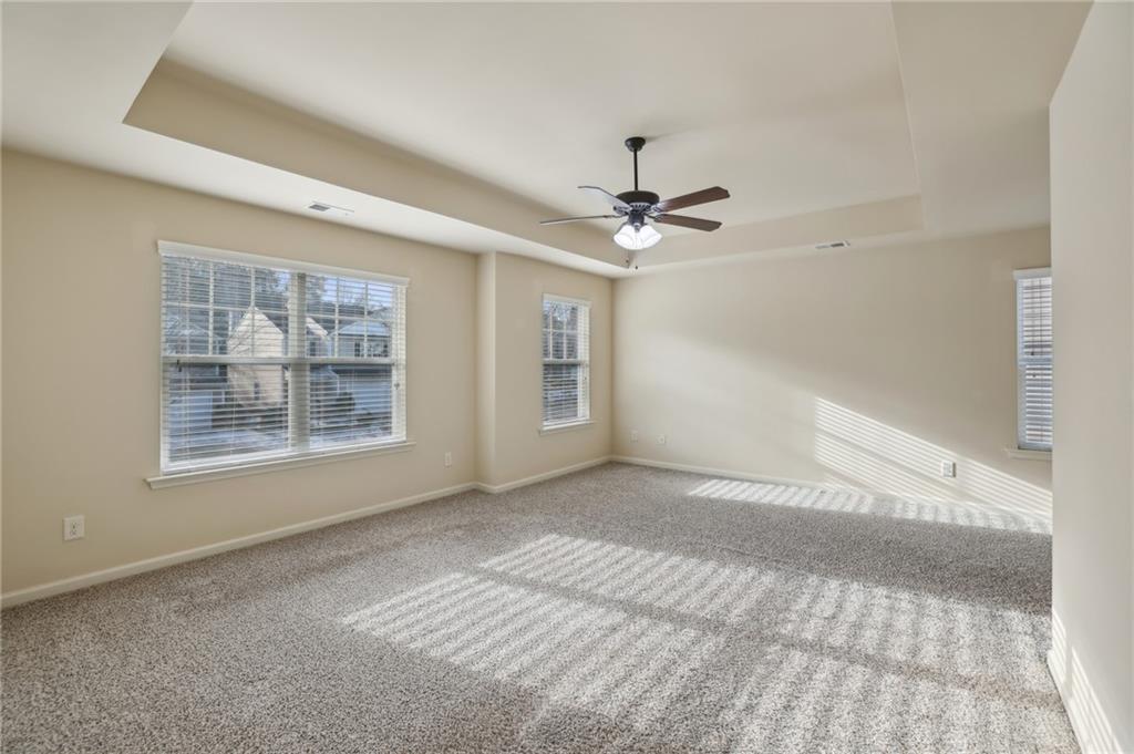 Empty bedroom with gray carpet, a tray ceiling, and sunlight through three windows.
