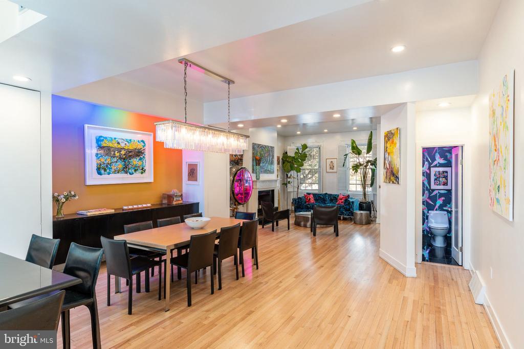 Open-plan dining room with a glass chandelier, colorful artwork, and a furnished sitting area beyond.