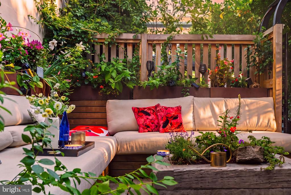 Garden patio with a cream sectional, red cushions, and lush plants.