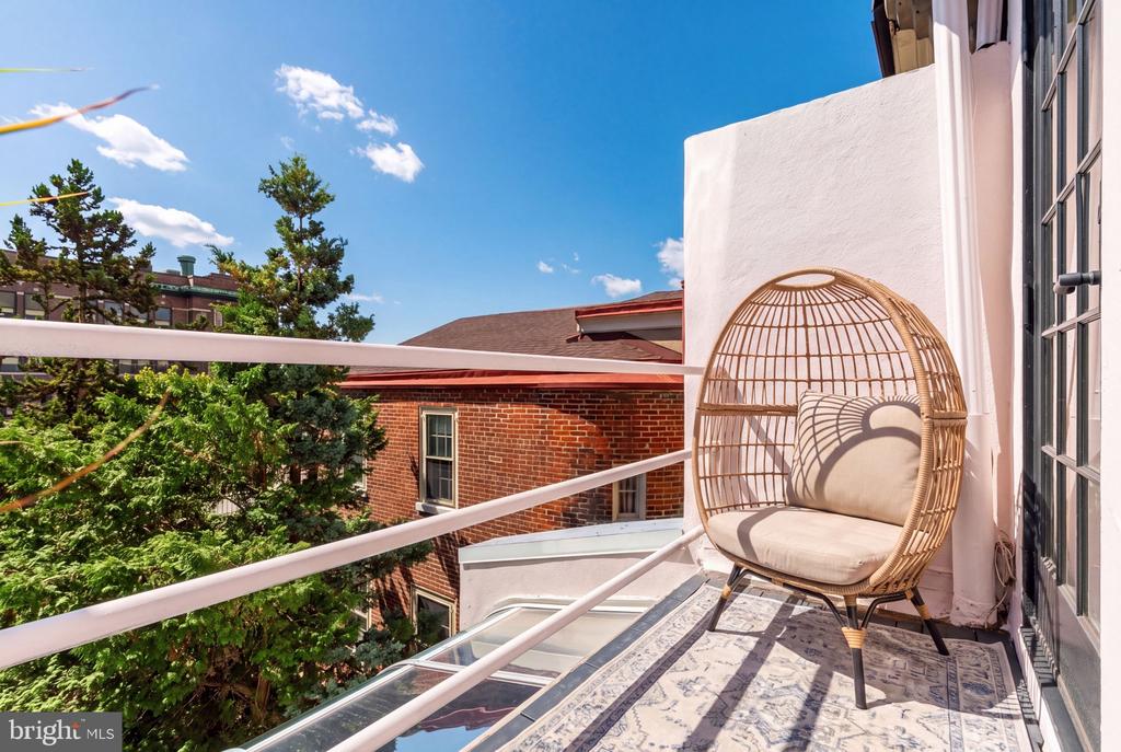 Balcony with a wicker egg chair and patterned rug overlooking trees and neighboring brick buildings.