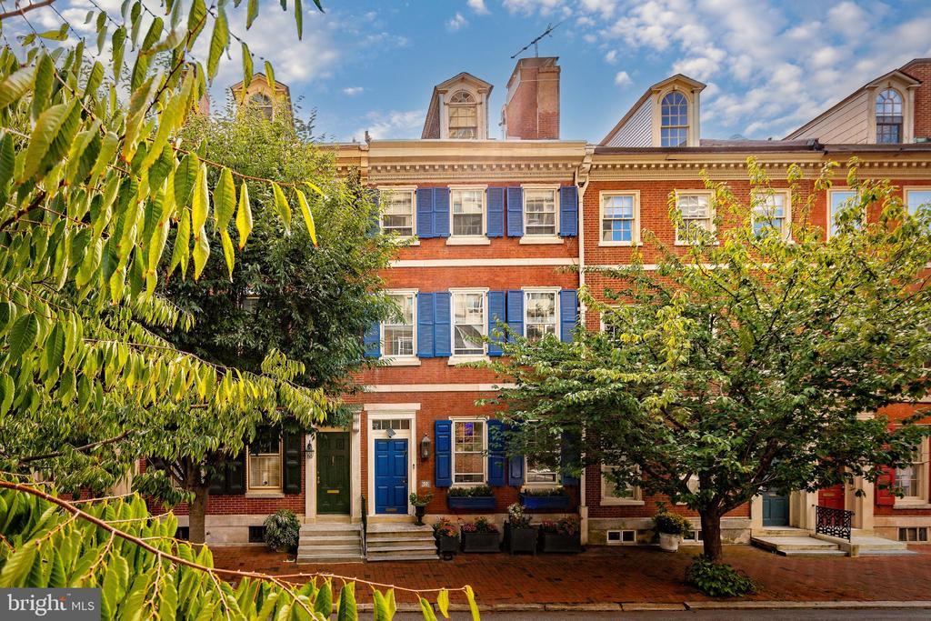 Red-brick Philadelphia row house with blue shutters and a blue door, framed by leafy trees.
