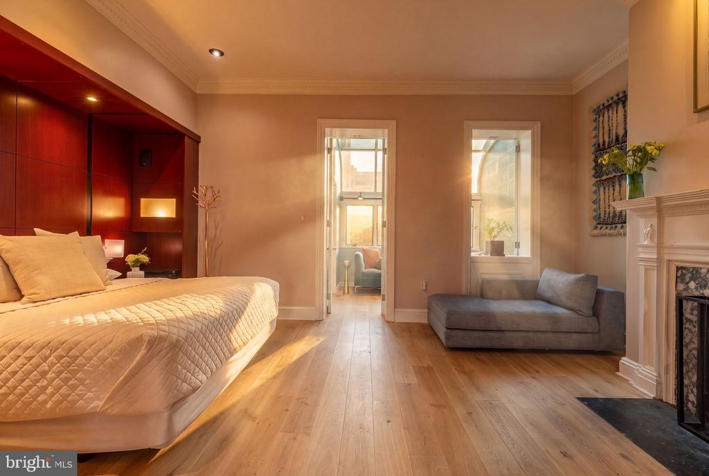 Furnished bedroom with dark wood bed wall, gray chaise lounge, fireplace, and tall windows.