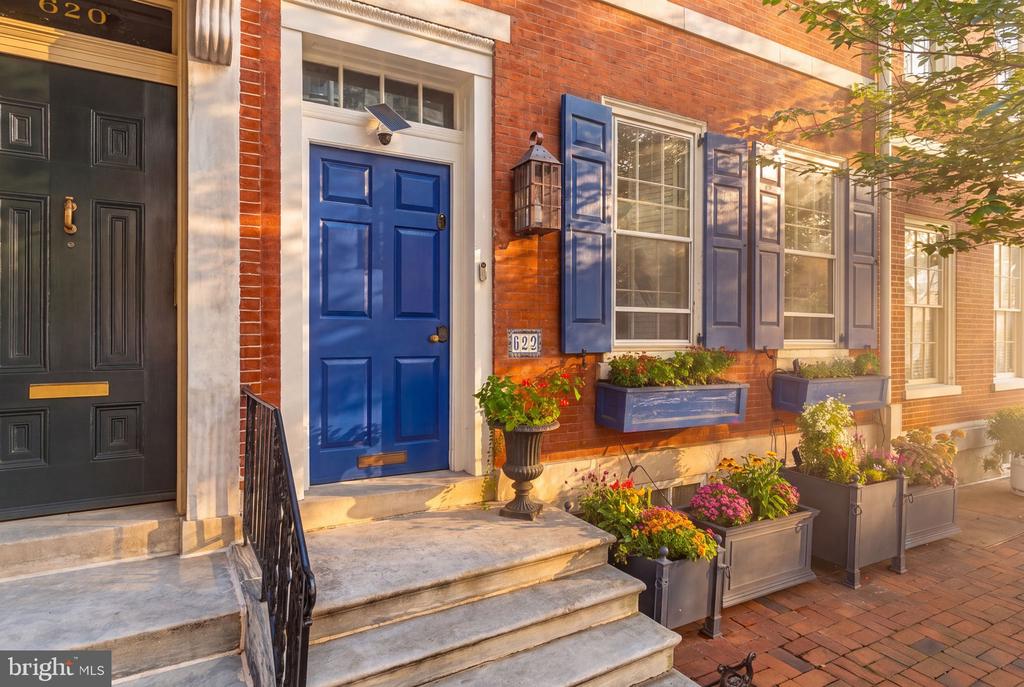 Red-brick rowhouse entrance with a blue door, stone steps, and flower-filled planters.