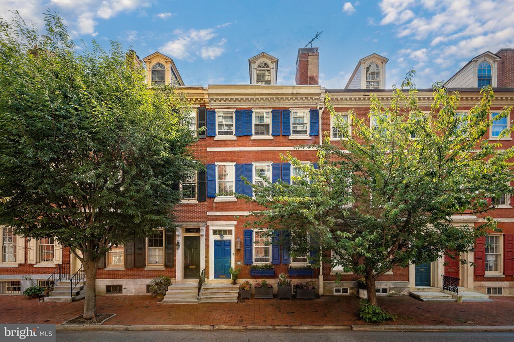 Red-brick rowhouse with blue shutters and dormer windows, partly framed by leafy trees.