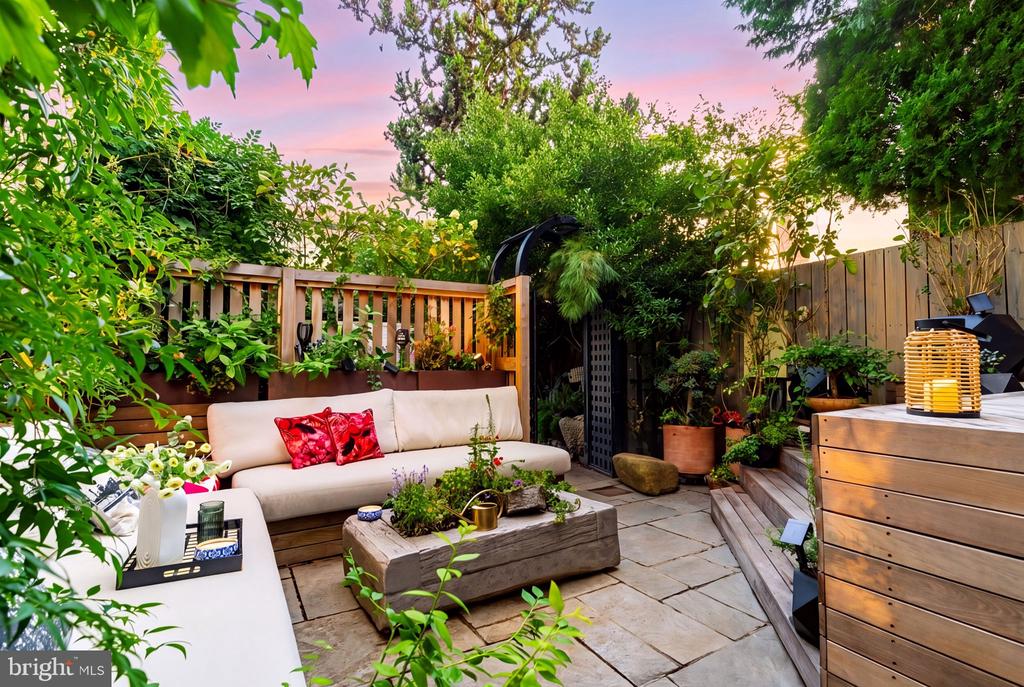 Furnished garden patio with stone paving, a cream sectional, and lush plants along a wooden fence.