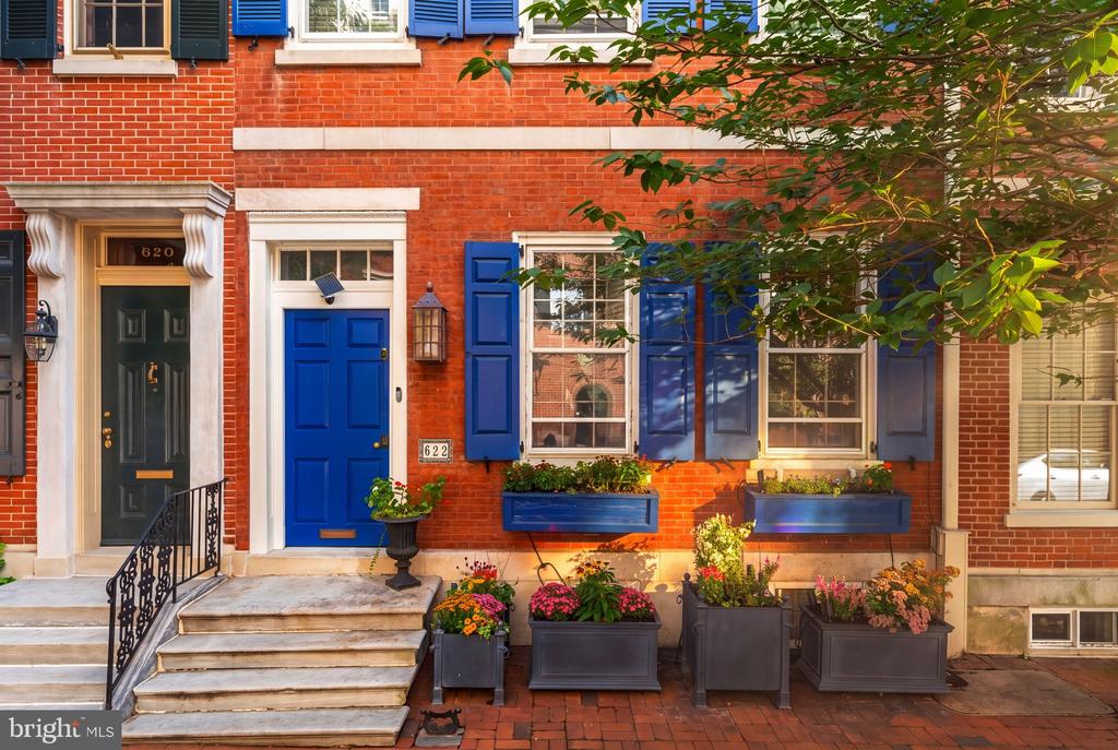 Red-brick row house with a bright blue door, blue shutters, stone steps, and flower planters.