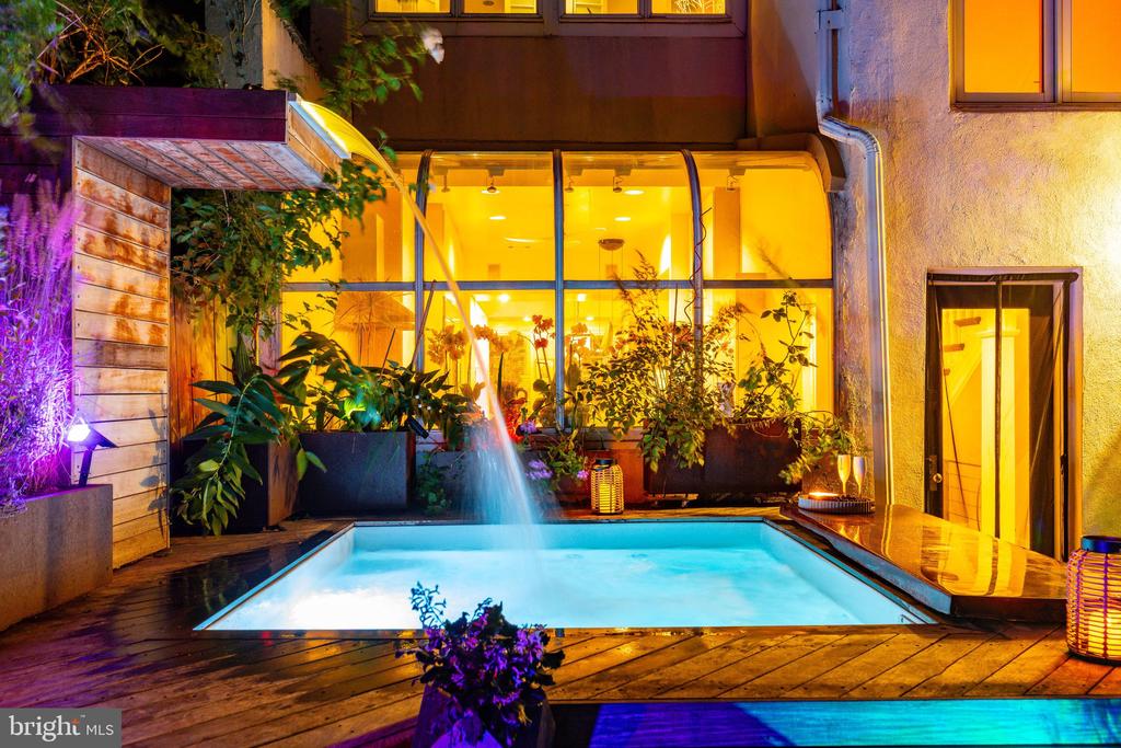 Courtyard pool with a fountain, surrounded by plants and warmly lit windows at night.
