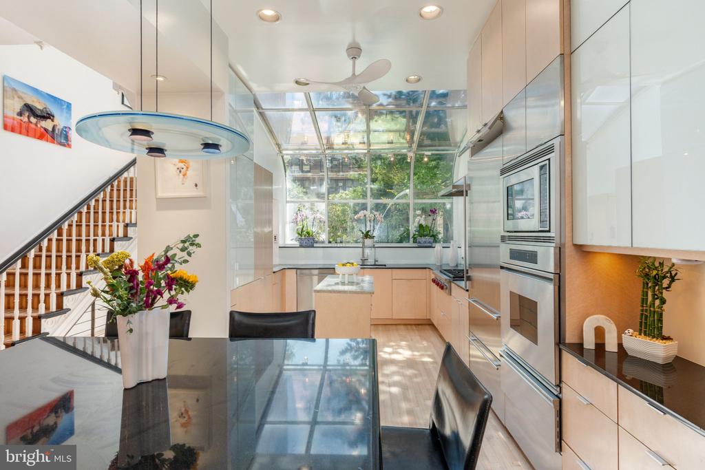 Modern kitchen with pale wood cabinetry, stainless appliances, and a glass ceiling overlooking greenery.