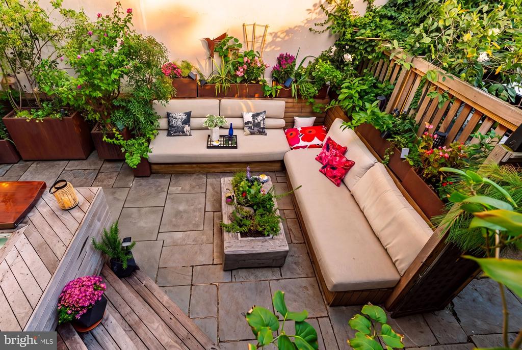 Outdoor patio with beige sectional seating, stone pavers, and abundant potted plants and flowers.