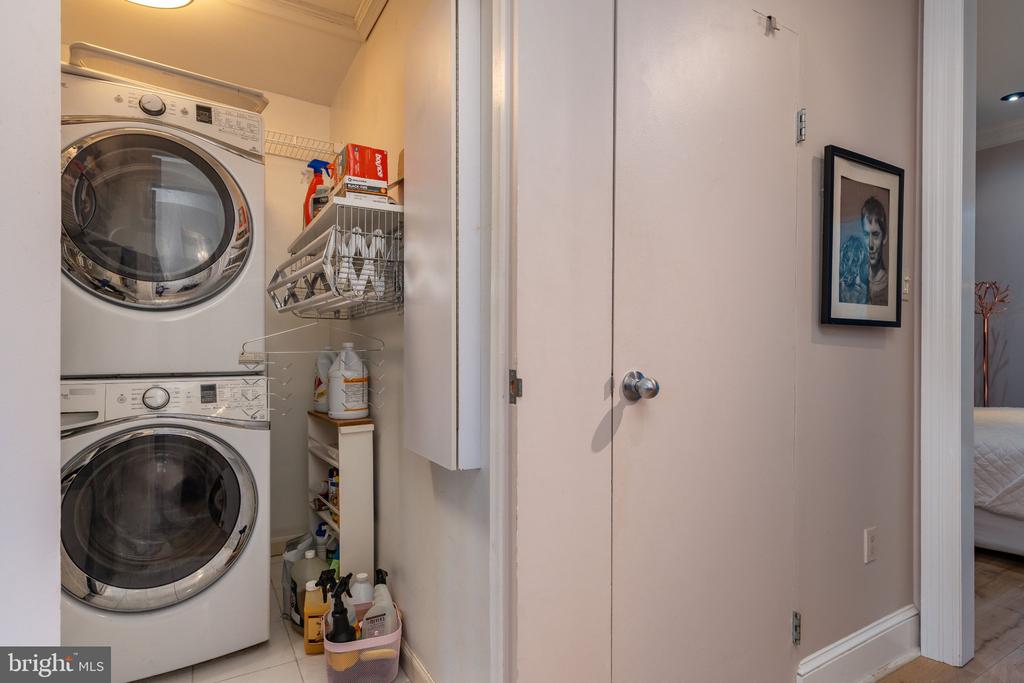 Laundry closet with stacked front-loading washer and dryer, open shelving, and cleaning supplies.