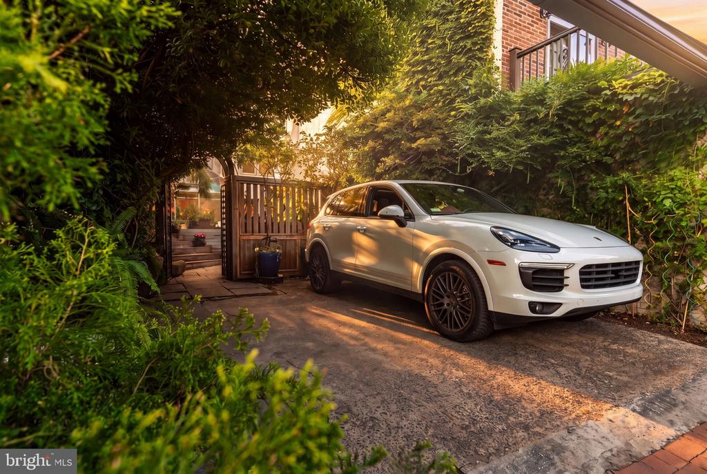 White SUV in a leafy driveway beside a brick home and wooden gate.